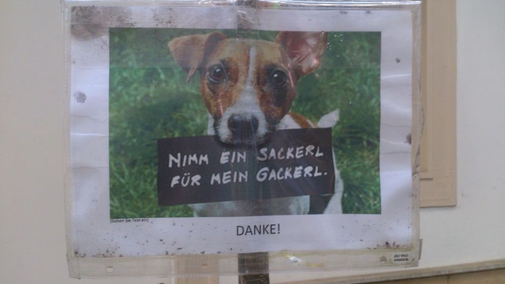 Hinweisschild, auf dem ein Hund mit einem Schild im Maul zu sehen ist. Auf dem Schild steht: "Nimm ein Sackerl für mein Gackerl."