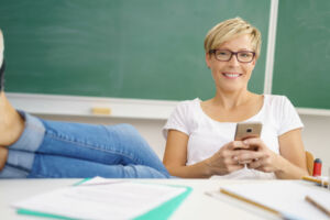 Eine junge Lehrerin sitzt mit einem Smartphone in der Hand am Schreibtisch vor der Tafel.