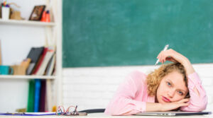 Junge Lehrerin sitzt erschöpft am Schreibtisch im Klassenzimmer.